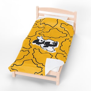 Op de afbeelding: Een gele deken met een zwart omtrekpatroon en een wit en zwart grafisch ontwerp op een houten bedframe. De deken is over een wit kussen en matras gedrapeerd. De afbeelding op de deken lijkt tekst te zijn.