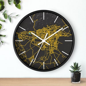 May include: A black wall clock with a white face and gold-colored city map design. The clock hands are white.