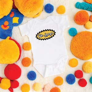 May include: White baby onesie with a yellow speech bubble that says "Durool  Ella" in black text.