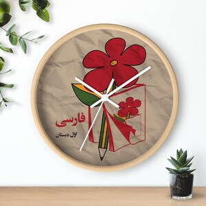 May include: A round clock with a brown background and a white clock face. The clock face features a red flower with a green stem and leaves, a yellow pencil, and a red book with a red flower on the cover. The text "قارسی" and "آذربایجان" is printed on the clock face.