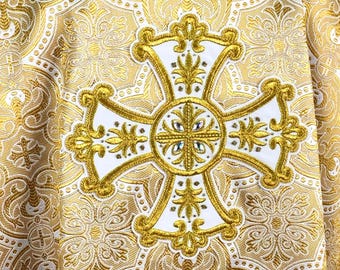 May include: Gold and white religious vestment with an ornate cross design. The vestment features a detailed pattern and a white collar. The cross is centered on the back and is outlined in gold.