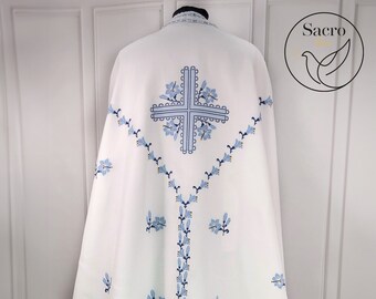May include: White liturgical vestment with blue embroidered flowers and a cross. The vestment has a decorative border around the bottom.