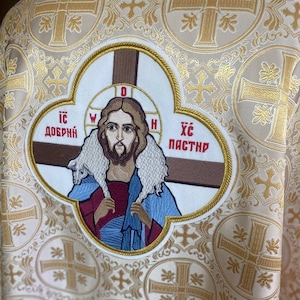 May include: A white fabric with a gold embroidered pattern of crosses and floral designs. A large embroidered patch features a depiction of Jesus Christ with two sheep, with the text "ИС ДОБРЫЙ" and "ХС ПАСТЫРЬ" in red.