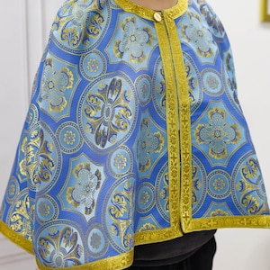 May include: A blue and gold patterned cape with a round collar and gold trim. The cape features a repeating floral design and fastens with a single gold button at the neck. The garment is made of a shiny fabric.