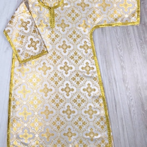 May include: A white liturgical vestment with a gold cross pattern and gold trim. The vestment has a round neckline and long sleeves.