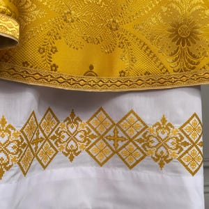 May include: A white liturgical vestment with a gold embroidered border. The border features a repeating pattern of diamonds and floral motifs.