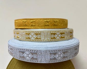May include: Three rolls of decorative trim, one gold, one white and gold, and one silver and white. The trim has a repeating pattern of stylized flowers and geometric shapes.