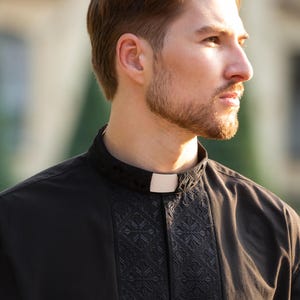 May include: A man wearing a black clergy shirt with a white collar and a black embroidered pattern. The shirt has a traditional clerical collar.