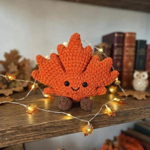 Puede incluir: Un juguete de peluche de hoja de arce naranja tejido a ganchillo con una cara sonriente, ojos de cuentas negras y dos pequeños pies marrones. La hoja texturizada tiene una forma distintiva de cinco puntas y detalladas puntadas de hilo.