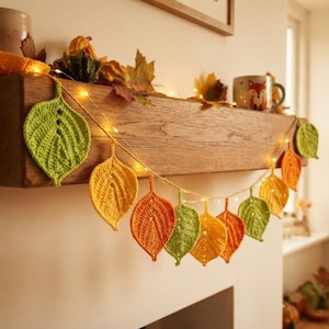 May include: A string of fairy lights featuring crocheted leaves in shades of green, yellow, and orange. The leaves are arranged along a thin wire, creating a decorative garland. The leaves have visible stitch patterns and pointed shapes.