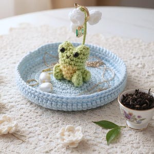May include: A light blue crocheted circular tray featuring a small green crocheted frog figure in the center. A tall, thin green stem rises from behind the frog, topped with white crocheted flowers. The tray holds two white beads and a gold chain.