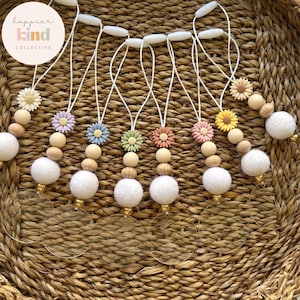 May include: Seven colorful flower charm necklaces with wooden beads, white pom poms, and clear acrylic circles. The necklaces are hanging on a woven basket.