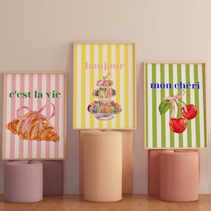 Könnte beinhalten: Drei gerahmte Drucke mit gestreiften Hintergründen und französischen Sätzen. Der erste Druck hat einen rosa-weiß gestreiften Hintergrund mit dem Satz "c'est la vie" und ein Croissant mit einem rosa Band. Der zweite Druck hat einen gelb-weiß gestreiften Hintergrund mit dem Satz "bonjour" und einen Turm aus Macarons. Der dritte Druck hat einen grün-weiß gestreiften Hintergrund mit dem Satz "mon chéri" und zwei Kirschen mit einem rosa Band.