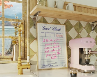 Puede incluir: Una máquina de espresso rosa con un vaso de café delante. Un cheque de invitado enmarcado con texto escrito a mano que dice "Creo que vamos a hacer una ensalada César, coca-cola dietética y papas fritas para la mesa" está colgado en la pared.