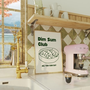 Op de afbeelding: Een keukeninterieur met een roze espressomachine, een ingelijste poster met de tekst "Dim Sum Club" en een glas koffie. Een gouden kraan en een raam met uitzicht op een meer en herfstbladeren zijn ook zichtbaar.