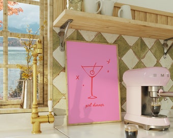 May include: A pink framed print with a red illustration of a martini glass and the text "girl dinner". The print is hanging on a wall with green and white diamond patterned tiles. A pink espresso machine is on the counter below the print.