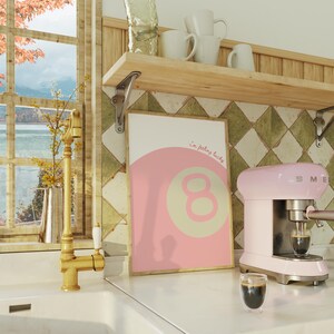 May include: A pink and beige poster with the text "i'm feeling lucky" and a large number eight in the center. The poster is hanging on a wall with green and white tile. A pink espresso machine is on the counter next to a glass of coffee.