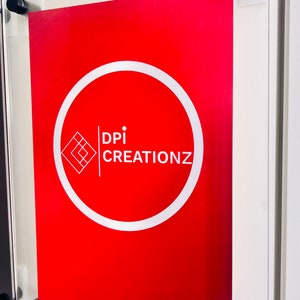 May include: A red sign with a white circle and the text "DPI CREATIONZ" inside the circle. The sign is framed in a clear acrylic frame with black mounting hardware.