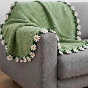 Puede incluir: Una manta verde tejida a ganchillo sobre un sofá gris. La manta está bordeada con flores de margarita blancas y amarillas con hojas verdes. La manta está hecha de un hilo suave y texturizado.