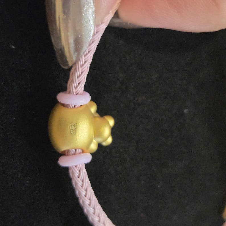 K&ouml;nnte beinhalten: Nahaufnahme eines goldfarbenen Charms mit Schweinchen-Design, befestigt an einem rosa geflochtenen Band. Der Charm hat einen glatten, abgerundeten K&ouml;rper und einen detaillierten Schweinekopf. Zwei rosa Ringe befinden sich auf beiden Seiten des Charms. Das Band wird von einer Person gehalten.