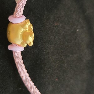 K&ouml;nnte beinhalten: Nahaufnahme eines goldfarbenen Anh&auml;ngers mit detailliertem Design, verbunden mit einem rosa geflochtenen Band. Der Anh&auml;nger wird von zwei hellrosa Ringen flankiert. Band und Anh&auml;nger vor dunklem Hintergrund.