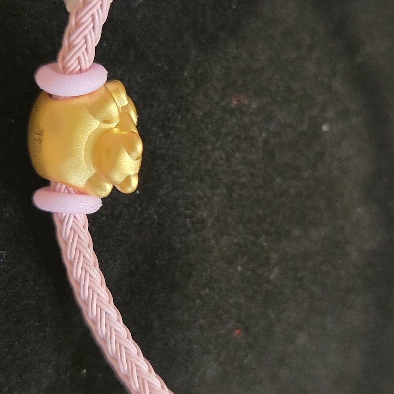 K&ouml;nnte beinhalten: Nahaufnahme eines goldfarbenen Anh&auml;ngers mit detailliertem Design, verbunden mit einem rosa geflochtenen Band. Der Anh&auml;nger wird von zwei hellrosa Ringen flankiert. Band und Anh&auml;nger vor dunklem Hintergrund.