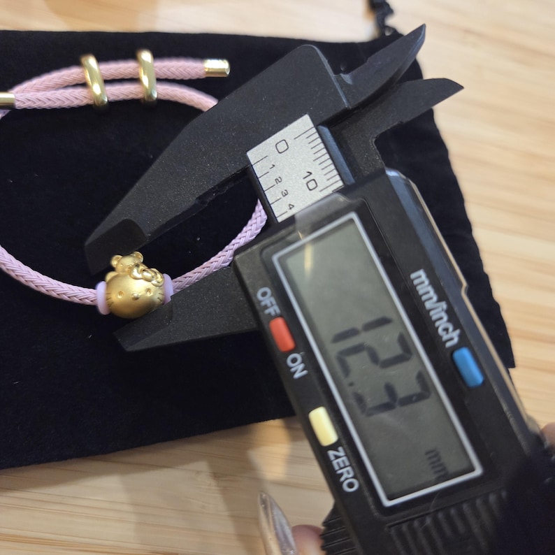 K&ouml;nnte beinhalten: Ein goldfarbenes Armband mit einem rosa Band. Der Anh&auml;nger ist ein Charakterdesign. Das Armband wird mit einem digitalen Messschieber auf 1,23 cm gemessen.