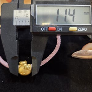 K&ouml;nnte beinhalten: Ein goldfarbener Hello Kitty-Anh&auml;nger wird mit einem digitalen Messschieber gemessen. Der Anh&auml;nger ist an einem rosa geflochtenen Band befestigt. Der Messschieber zeigt 1,14 mm an.