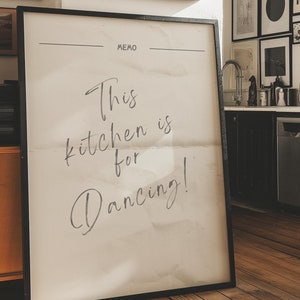 May include: A black framed poster with the text "This kitchen is for dancing!" on a white background. The poster is hanging on a wall in a kitchen with a wooden floor.