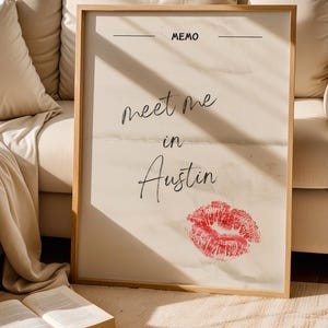 May include: A framed print with the words "meet me in Austin" in cursive script, a red lipstick kiss mark, and the word "MEMO" at the top. The frame is light wood.