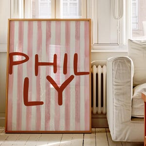 May include: A framed art print featuring the word "PHILLY" in a bold, rust-colored font. The background has vertical stripes in alternating shades of pink and off-white. The frame is a light wood color.