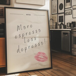 May include: A black framed poster with the text "More espresso less depresso!" and a pink lipstick kiss mark on a white background. The poster is hanging on a wall in a kitchen.