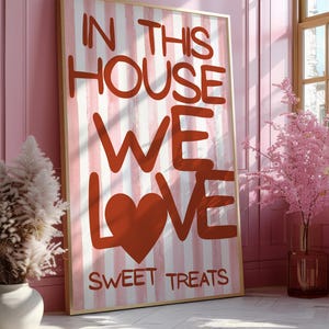 Könnte beinhalten: Ein gerahmter Kunstdruck mit goldenem Rahmen, der an einer rosa Wand lehnt. Der Druck zeigt rote und weiße vertikale Streifen und die Worte "IN THIS HOUSE WE LOVE SWEET TREATS" in roten Blockbuchstaben. Dekorative Elemente sind eine Vase mit Pampasgras und ein rosa Blumenarrangement.