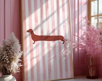 May include: A framed art print featuring a dachshund dog illustration. The artwork has a brown dachshund on a pink and white striped background. The frame is gold. The print is displayed in a pink room with a window and decorative plants.