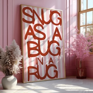 Puede incluir: Una impresión artística enmarcada con la frase "SNUG AS A BUG IN A RUG" en letras rojas sobre un fondo a rayas rosas y blancas. El marco es dorado. La obra de arte se exhibe en una habitación rosa con plantas decorativas.