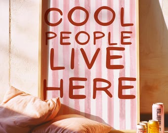 May include: A framed print with the text "COOL PEOPLE LIVE HERE" in a bold, red font against a pink and white striped background. Two peach-colored pillows rest on the floor, along with several cans of beer and a magazine.