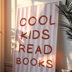 Peut inclure: Affiche encadrée avec le texte "COOL KIDS READ BOOKS" en lettres rouille. Le fond présente des rayures verticales blanches et roses claires. L'affiche est appuyée contre un mur de briques blanches.