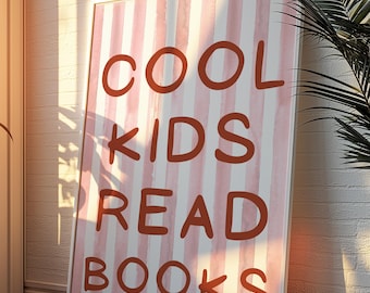 May include: A framed poster with the text "COOL KIDS READ BOOKS" in a bold, rust-coloured font. The background features vertical stripes in white and light pink. The poster is leaning against a white brick wall.