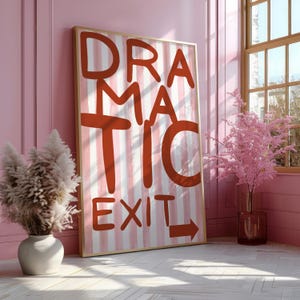 Puede incluir: Una impresión artística enmarcada con las palabras "DRAMATIC EXIT" en letras rojas sobre un fondo a rayas. La obra de arte está apoyada contra una pared rosa, con un jarrón de hierba de la pampa y una ventana con un árbol rosa en el fondo.