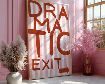 May include: A framed art print with the words "DRAMATIC EXIT" in red letters on a striped background. The artwork is leaning against a pink wall, with a vase of pampas grass and a window with a pink tree in the background.