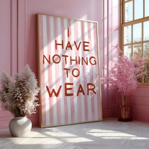 Puede incluir: Un cuadro enmarcado con el texto "I HAVE NOTHING TO WEAR" en letras rojas sobre un fondo de rayas rosas y blancas. El marco dorado está apoyado contra una pared rosa. Un jarrón con flores secas está a la izquierda.