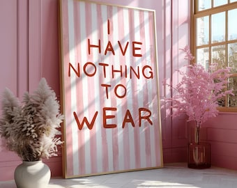May include: A framed art print with the text "I HAVE NOTHING TO WEAR" in red letters against a pink and white striped background. The frame is gold, and the artwork is leaning against a pink wall. A vase with dried flowers is on the left.