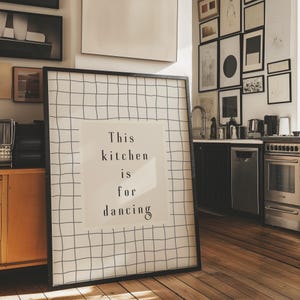 Puede incluir: Un estampado enmarcado con un patrón de cuadrícula negra y el texto "This kitchen is for dancing" en un entorno de cocina. El estampado está apoyado contra una pared con varias obras de arte enmarcadas. La cocina cuenta con pisos de madera, un horno de acero inoxidable y gabinetes negros.