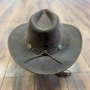 May include: A brown leather cowboy hat with a wide brim and a rounded crown. The hat features a decorative band with silver-colored floral accents and a chin strap. The hat is made of distressed leather.