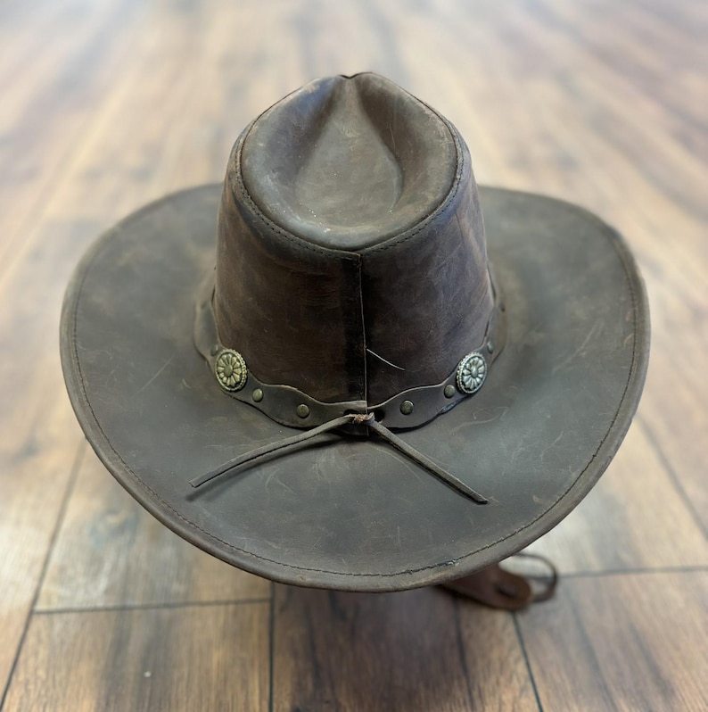 May include: A brown leather cowboy hat with a wide brim and a rounded crown. The hat features a decorative band with silver-colored floral accents and a chin strap. The hat is made of distressed leather.