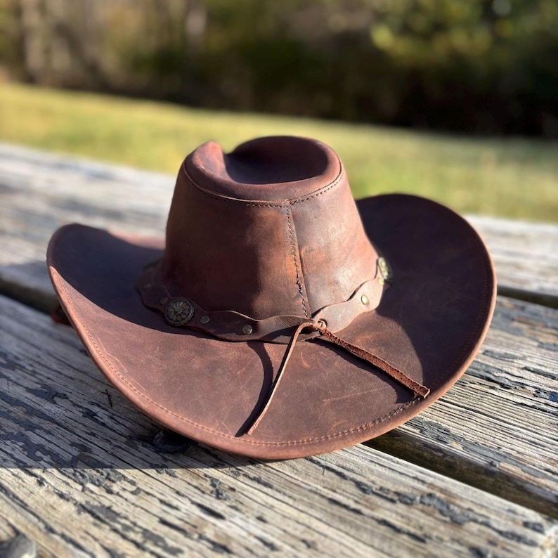 May include: A brown leather cowboy hat with a wide brim and a rounded crown. The hat features a decorative band with metal accents and a leather chin strap. The hat is resting on a wooden surface.