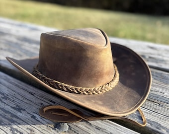 May include: A brown leather cowboy hat with a braided leather band. The hat has a distressed look and is lying on a wooden surface.