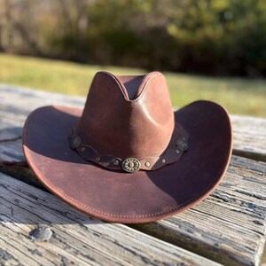 May include: A brown leather cowboy hat with a decorative band featuring floral accents. The hat has a classic brim and crown design, suitable for outdoor wear. The hat is resting on a weathered wooden surface.
