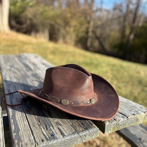May include: A brown leather cowboy hat with a wide brim and a decorative band featuring metal embellishments. The hat is resting on a weathered wooden surface, with a natural outdoor background.