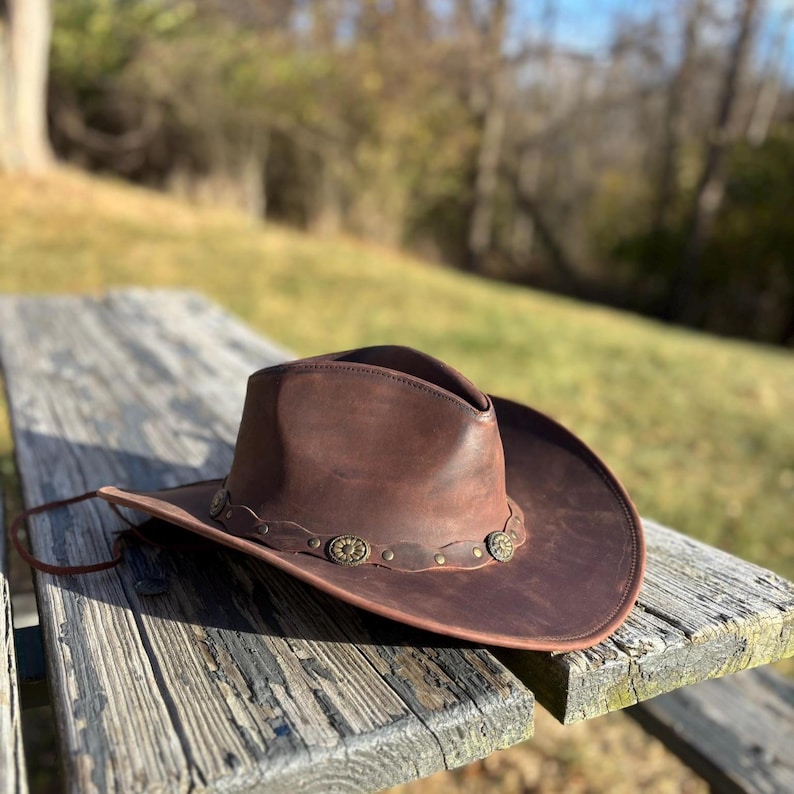 May include: A brown leather cowboy hat with a wide brim and a decorative band featuring metal embellishments. The hat is resting on a weathered wooden surface, with a natural outdoor background.